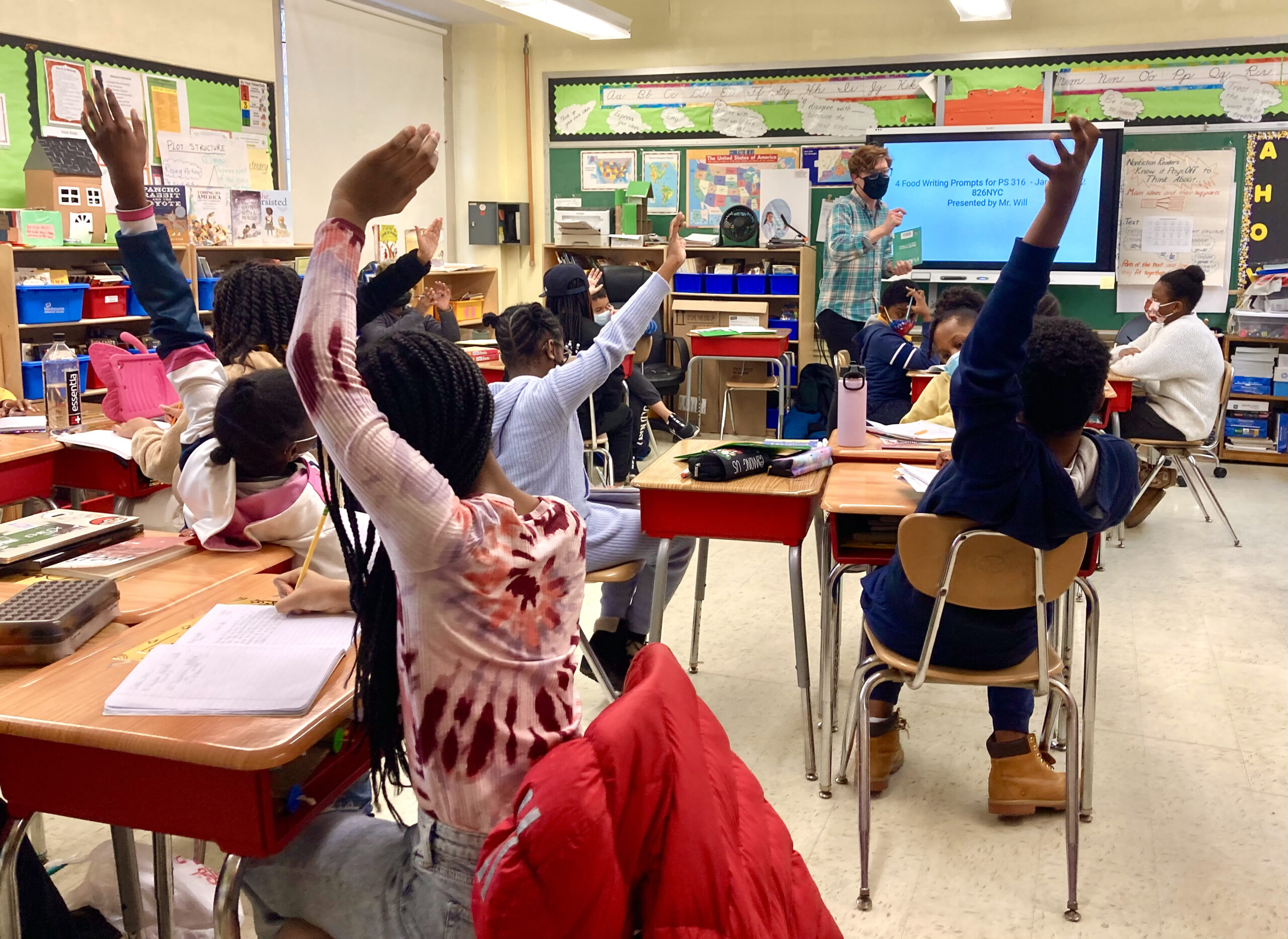 Los estudiantes en un salón de clases de Young Writers Publish levantan la mano con entusiasmo.
