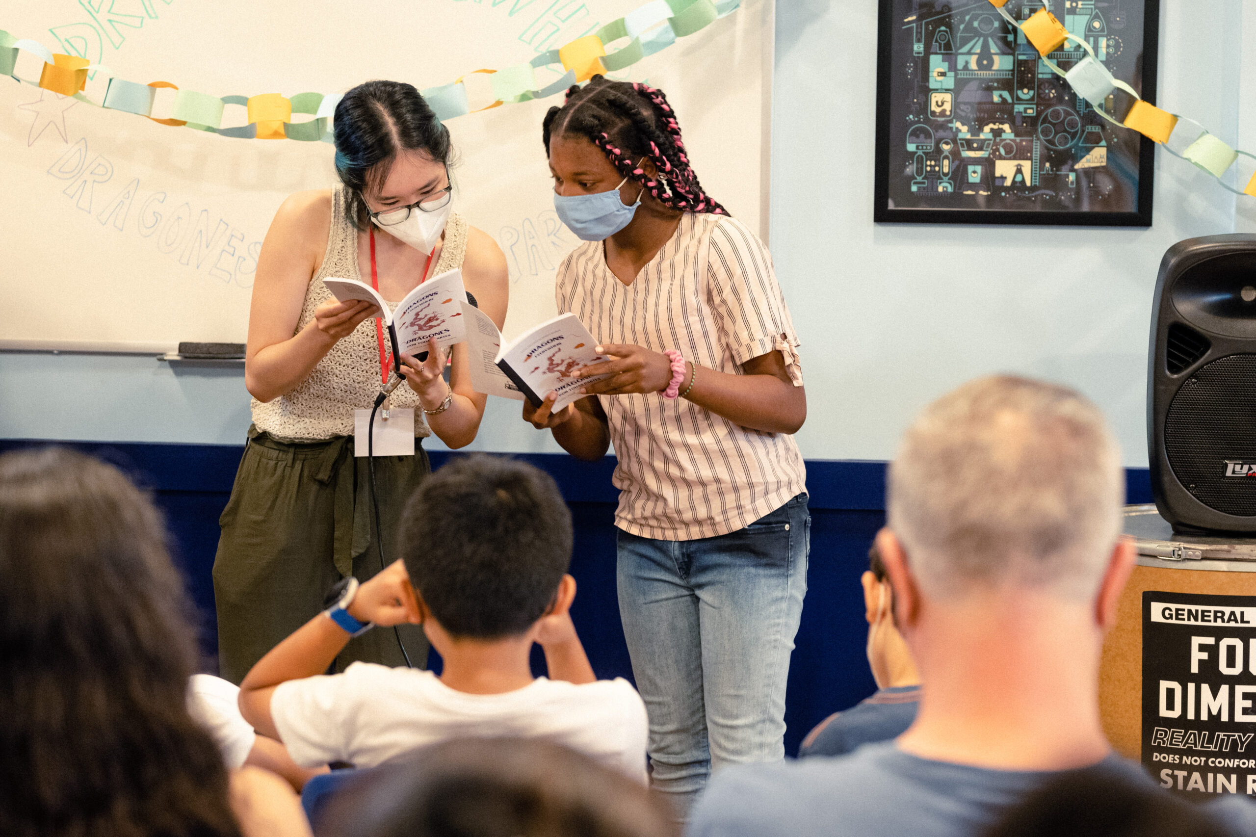 Un estudiante y un miembro del personal miran juntos copias de un libro de notas para estudiantes, frente a una audiencia que espera.