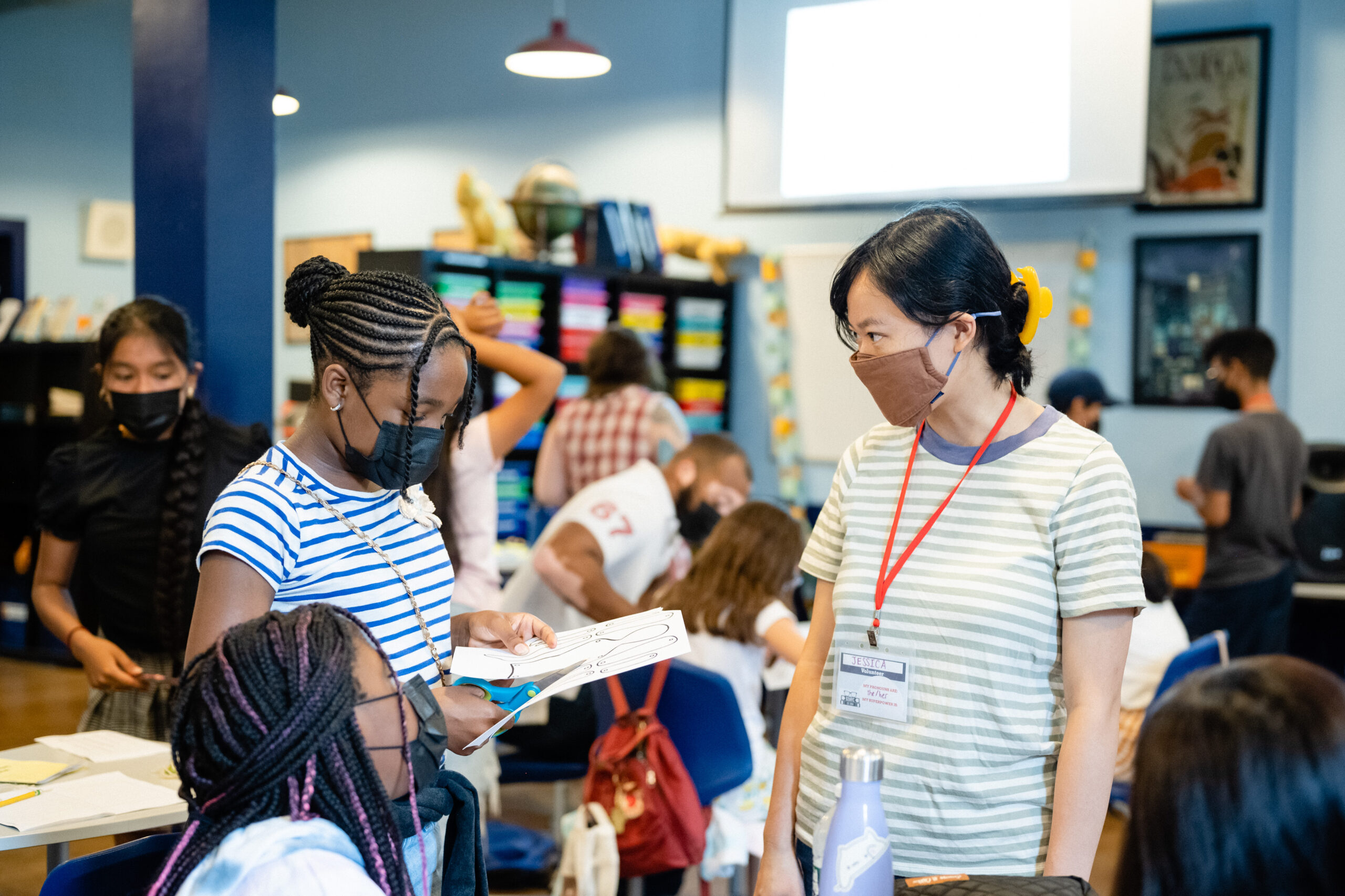 An 826NYC volunteer with a happy young author at our youth writing center. Both the volunteer and student are standing and other young authors and adult volunteers are visible in the foreground and background.