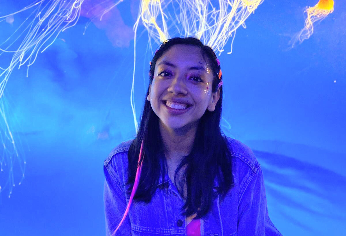 January volunteer of the month, Stephanie Frescas, standing in front of a jellyfish tank. She is smiling and the jellyfish are floating above her head.