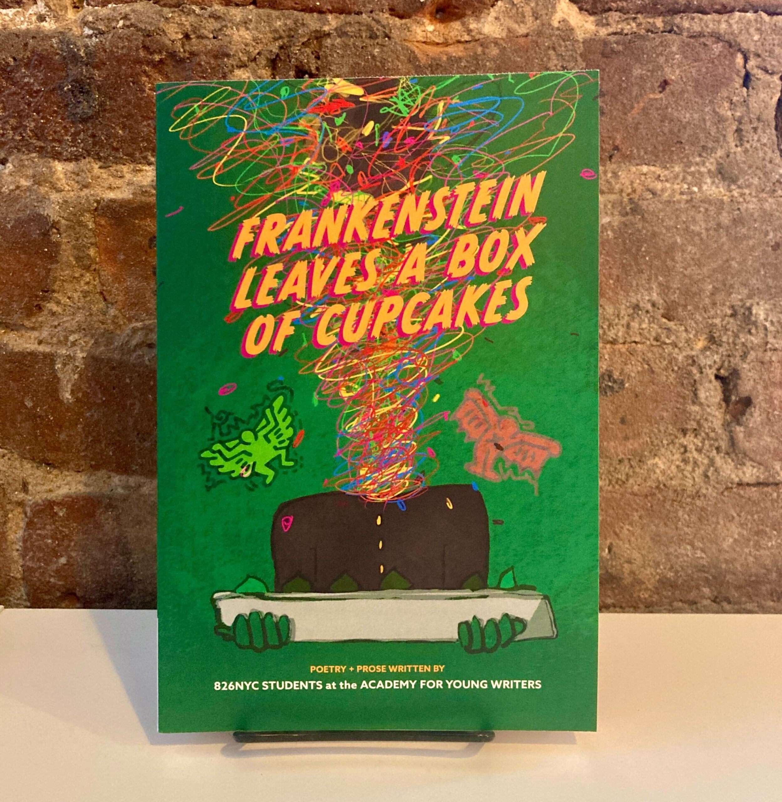 Portada de Frankenstein Leaves a Box of Cupcakes, un libro de bolsillo. El título está en letras naranjas vibrantes sobre un fondo verde, con una figura cuya cabeza es un garabato gigante que sostiene una bandeja.