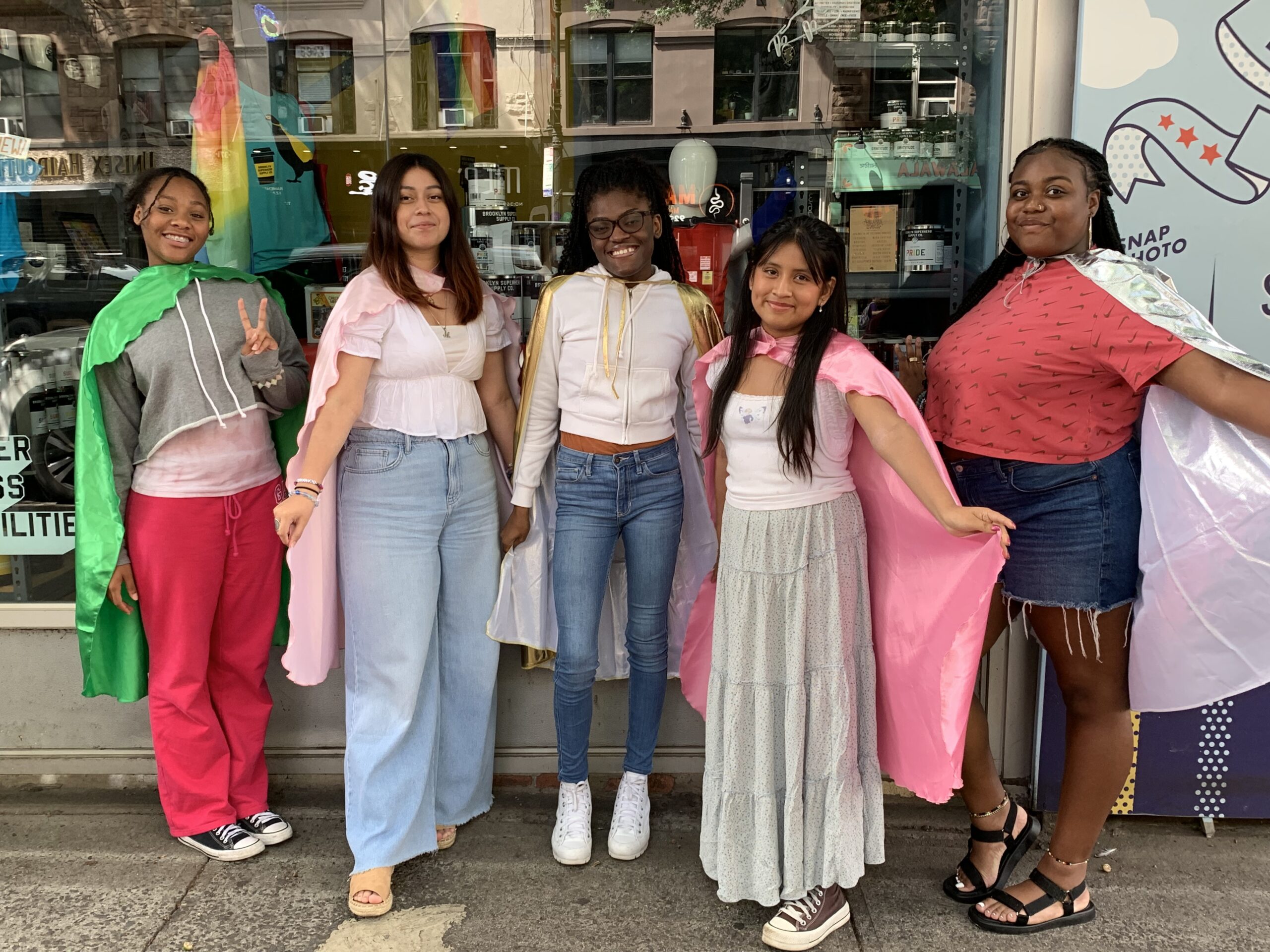 Five teen interns wearing capes and standing in front of 826NYC. From left to right: Kayla, Mayra, Jordan, Adalia, and Aleyah
