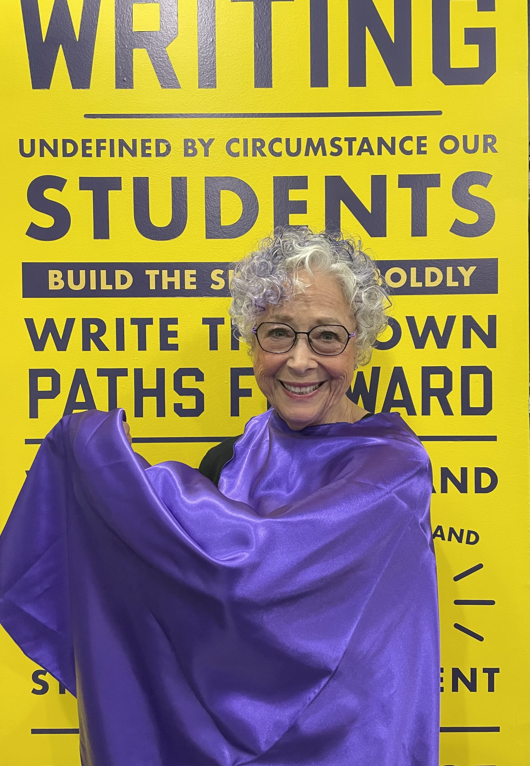 826NYC volunteer Barbara standing in front of a yellow background, wearing a purple cape and smiling. She has gray hair with a purple streak and black glasses