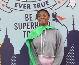 826NYC Student Kayla Sabb, wearing a shiny green cape in front of a light blue background