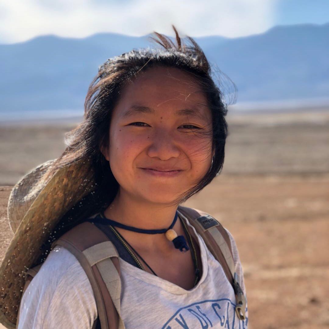 826NYC volunteer Brook standing wearing a white t-shirt, a gray backpack, and a brimmed hat tied around her neck. She is smiling and her dark brown hair is being blown by the wind. There is sand and mountains behind her.