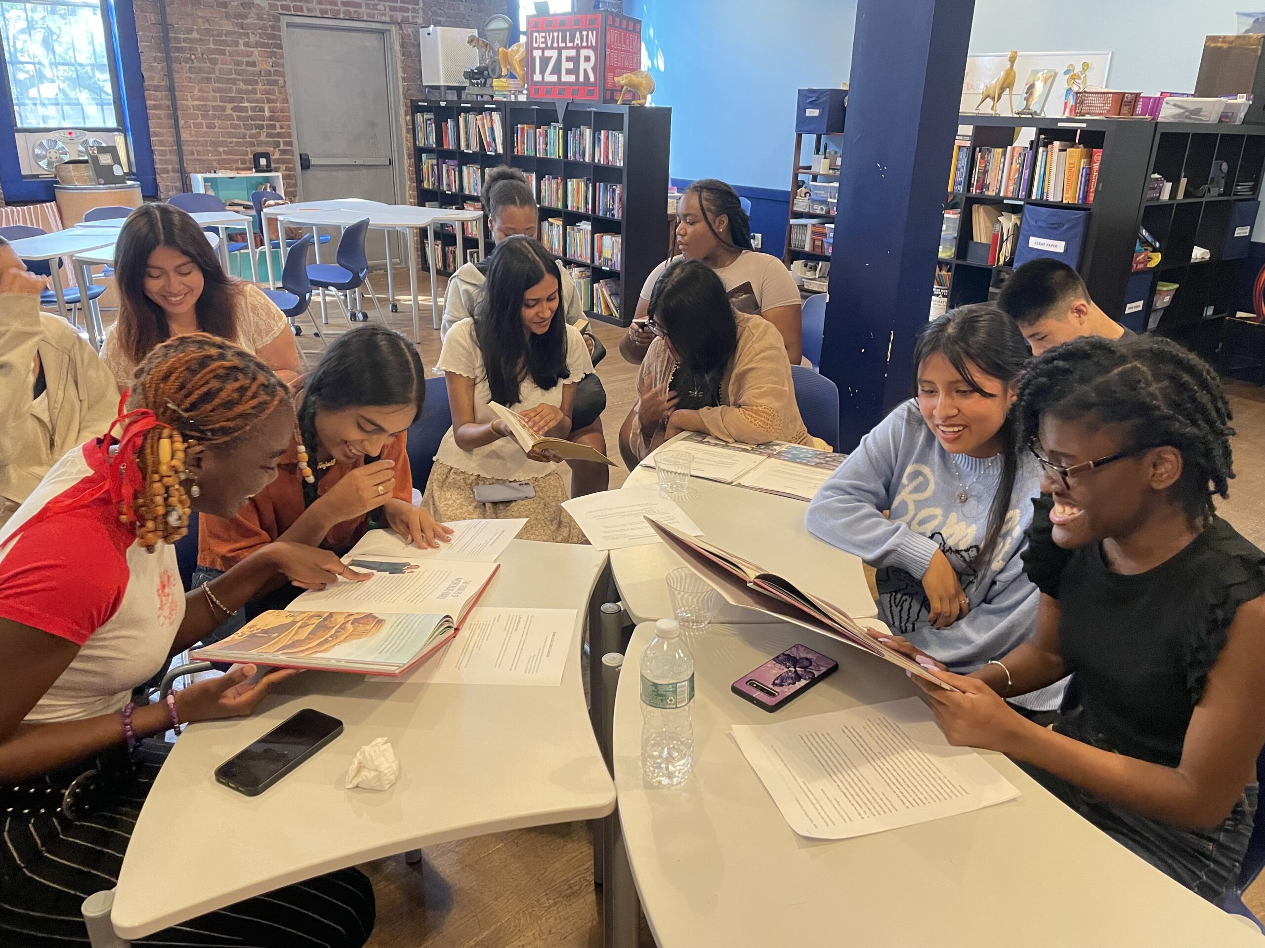 A group of teen writers sits at a cluster of tables happily reading and writing together.