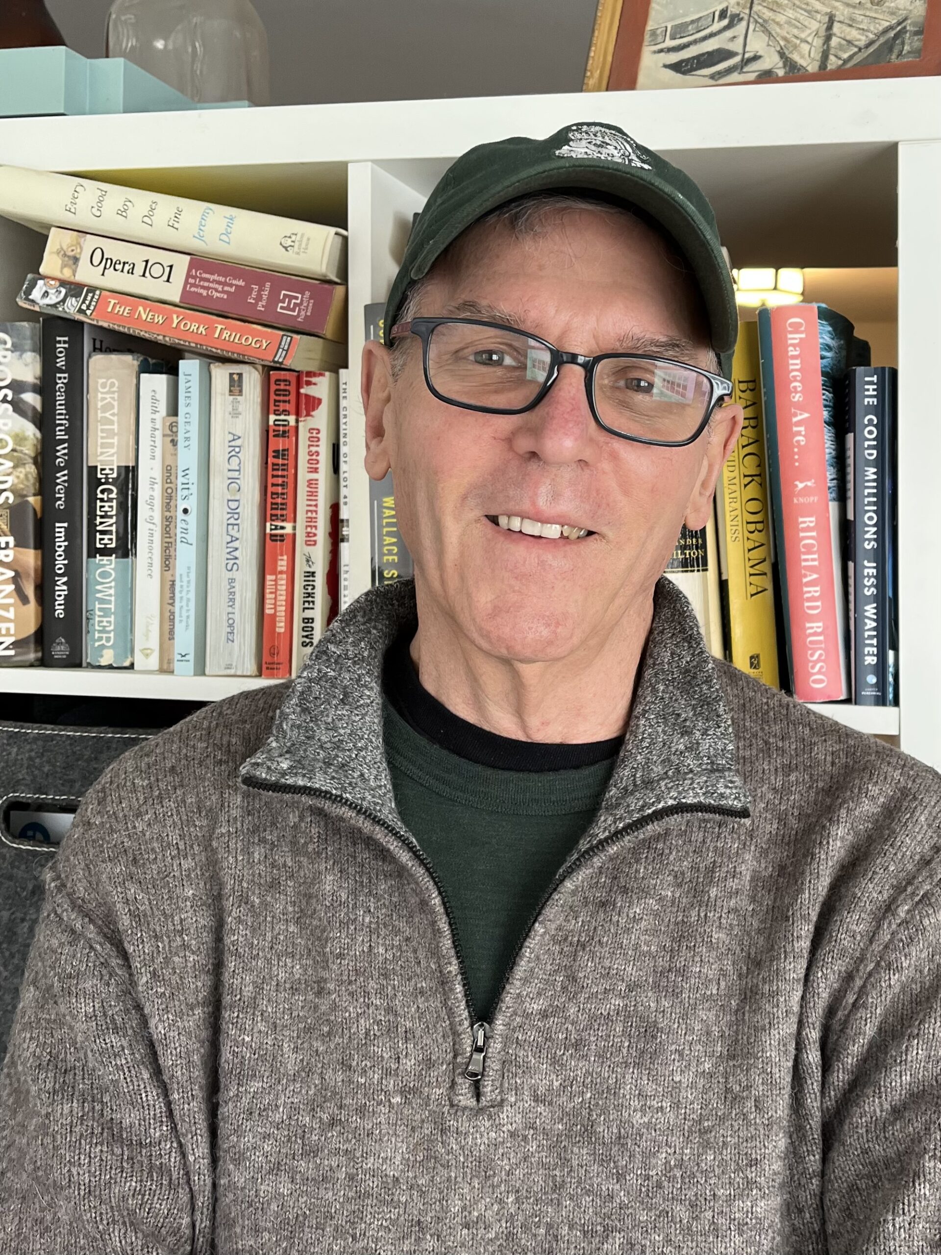 El voluntario Steve Giegerich lleva gafas negras cuadradas, una gorra de béisbol negra y un suéter gris. Está parado frente a una estantería y sonriendo.