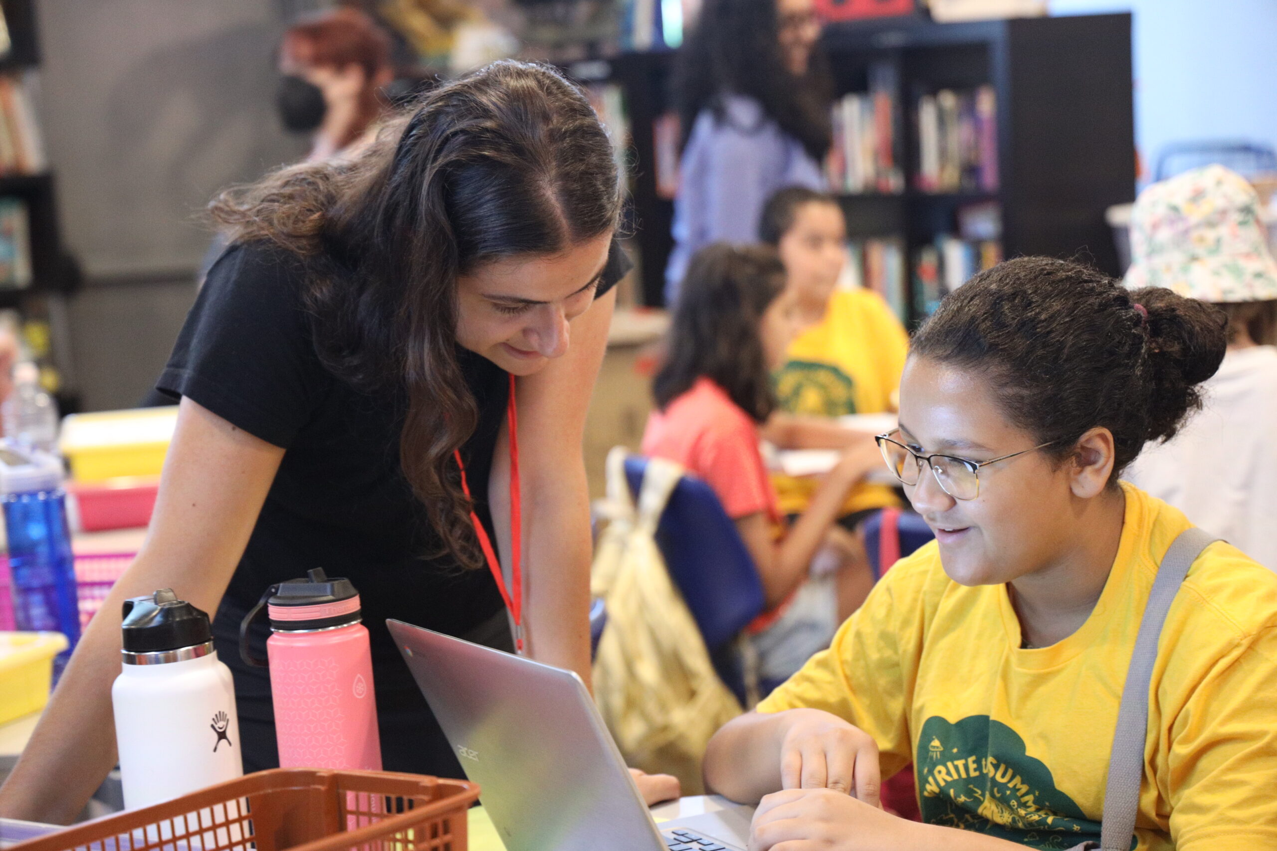 A student smiles as they type up a story and a volunteer looks on encouragingly.